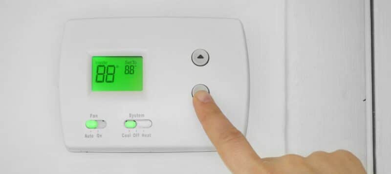 A finger is about to press a button on a white digital thermostat displaying