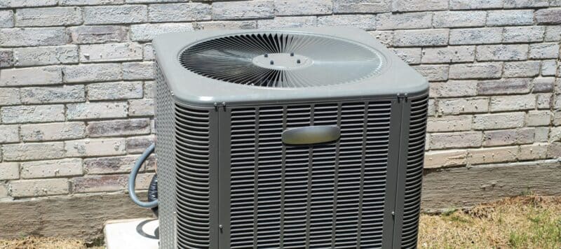An outdoor air conditioning unit sits on a concrete pad next to a brick wall, surrounded by sparse grass