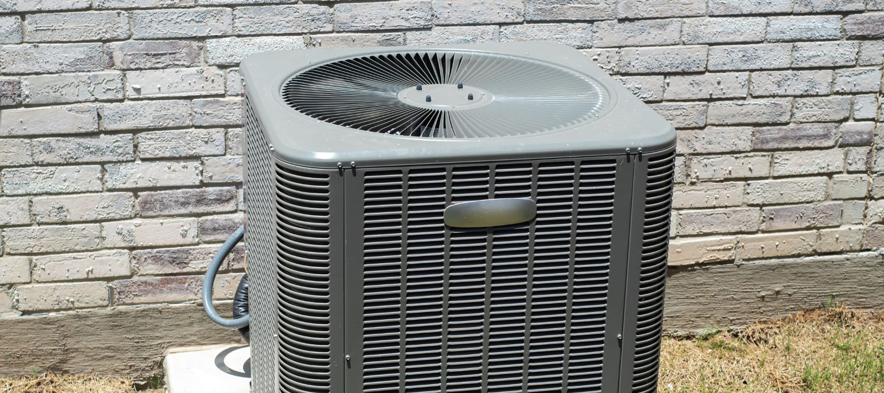 An outdoor air conditioning unit sits on a concrete pad next to a brick wall, surrounded by sparse grass