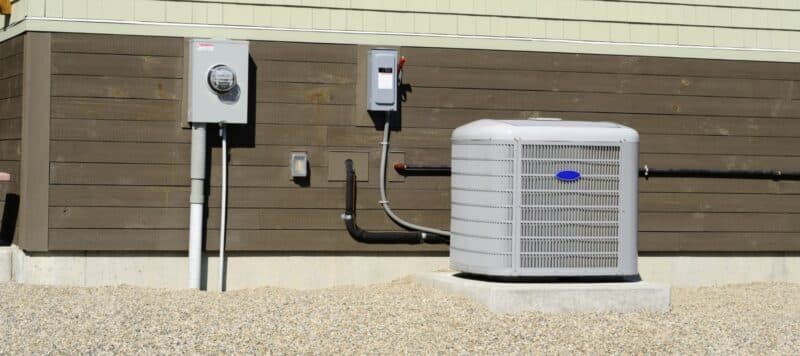 Outdoor air conditioning unit installed beside a brown house with utility boxes and piping