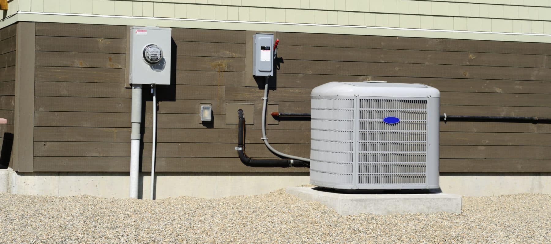 Outdoor air conditioning unit installed beside a brown house with utility boxes and piping