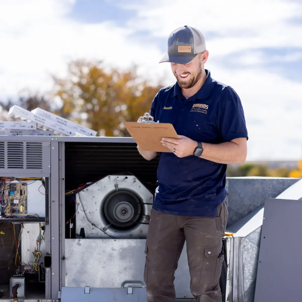 A smiling technician in a blue polo and cap holds a clipboard while inspecting rooftop HVAC equipment on a sunny day with trees in the background.