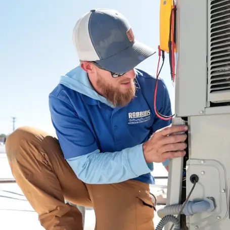 A technician wearing a blue Robbins shirt and cap kneels while inspecting and repairing an outdoor air conditioning unit with tools and wires attached.