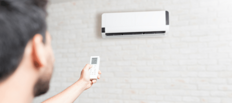 A person holding a remote control and pointing it at a wall-mounted air conditioner unit in a room with a light-colored brick wall.