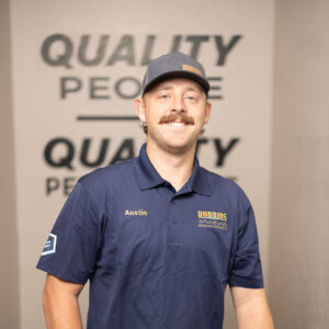 A man with a mustache wearing a navy blue polo shirt and cap stands indoors, smiling with one hand resting on HVAC equipment. The shirt has Austin and Robbins Heating & Air Conditioning logos. “QUALITY PEOPLE” is on the wall behind him.
