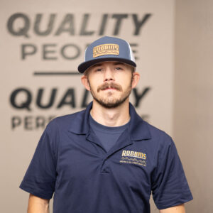 A man with a mustache and goatee wearing a blue Robbins Heating & Air Conditioning polo and matching hat stands indoors, posing with one hand in his pocket. The words QUALITY PEOPLE appear blurred in the background.