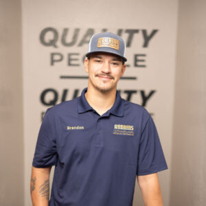 A man wearing a blue Robbins Service polo and cap stands indoors, smiling with one hand in his pocket and the other resting on HVAC equipment. The wall behind him reads QUALITY PEOPLE.