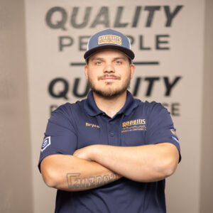 A man wearing a blue Robbins Heating & Air Conditioning shirt and cap stands with arms crossed, smiling. The wall behind him has the words QUALITY PEOPLE partially visible. An air conditioning unit is nearby.