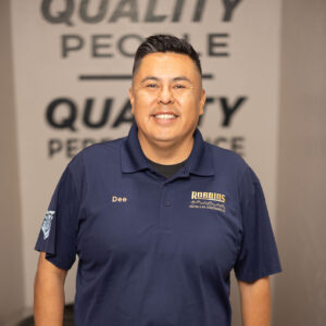 A man wearing a navy blue polo shirt with Dee and Robairs logos stands smiling indoors, resting one hand on an air conditioning unit. The background displays blurred QUALITY PEOPLE text on the wall.