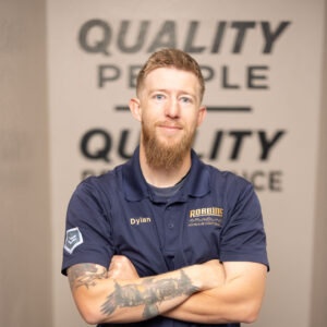 A man with a beard and tattoos, wearing a navy blue Robotic company shirt with the name Dylan, stands with arms crossed in front of a wall with the words QUALITY PEOPLE QUALITY SERVICE.