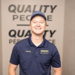 A smiling young man in a blue Robbins Heating & Air Conditioning polo and cap stands indoors with hands in pockets. The wall behind him reads QUALITY PEOPLE.