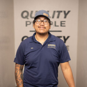A man wearing glasses, a cap, and a blue Robbins Heating & Air Conditioning polo stands indoors with one arm resting on HVAC equipment. He has tattoos on one arm and keys clipped to his belt.