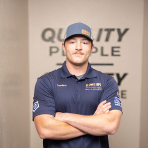A man wearing a blue cap and blue collared shirt with Isaiah and Rabbits embroidered on it stands with arms crossed in an indoor setting, in front of an HVAC unit. The words QUALITY PEOPLE are partially visible on the wall behind him.