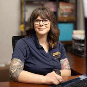 A woman with glasses and tattoos on her arms sits at an office desk, wearing a navy blue polo shirt. She is smiling and looking slightly past a computer monitor in front of her. Shelves are visible in the background.