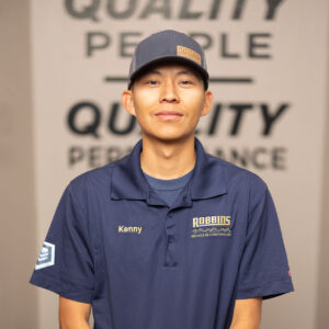A man wearing a navy blue Robbins polo shirt and cap stands indoors with hands clasped. The background features a blurred sign reading Quality People, Quality Performance.