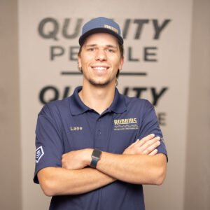 A man wearing a navy Robbins company polo shirt and cap stands with arms crossed, smiling in front of a blurred sign that reads QUALITY PEOPLE. He is indoors, near HVAC equipment.