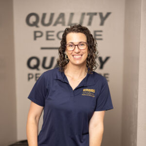 A smiling woman with curly hair and glasses, wearing a navy blue polo and jeans, stands indoors beside HVAC equipment. The words QUALITY PEOPLE are visible on the wall behind her.