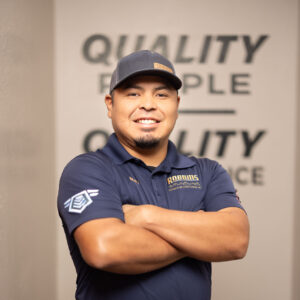 A man in a navy blue polo shirt and cap stands with arms crossed and smiles at the camera. The background features HVAC equipment and a wall with the words QUALITY PEOPLE QUALITY SERVICE.