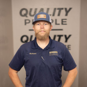 A man with a beard wearing a navy blue polo shirt and cap stands with hands in pockets in front of an HVAC unit. The shirt and cap have a Robbins logo, and Richard is embroidered on the shirt. A QUALITY PEOPLE sign is on the wall behind him.