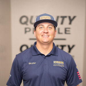A man wearing a navy blue polo shirt and matching cap, both branded Robbins, stands smiling with hands in his pockets. The name Skyler is embroidered on his shirt. The blurred background shows the word QUALITY.