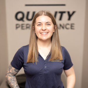 A woman with straight, light brown hair, wearing a navy polo shirt and light blue jeans, stands smiling with hands in her pockets. She has a visible arm tattoo and stands in front of a blurred sign reading QUALITY PERFORMANCE.