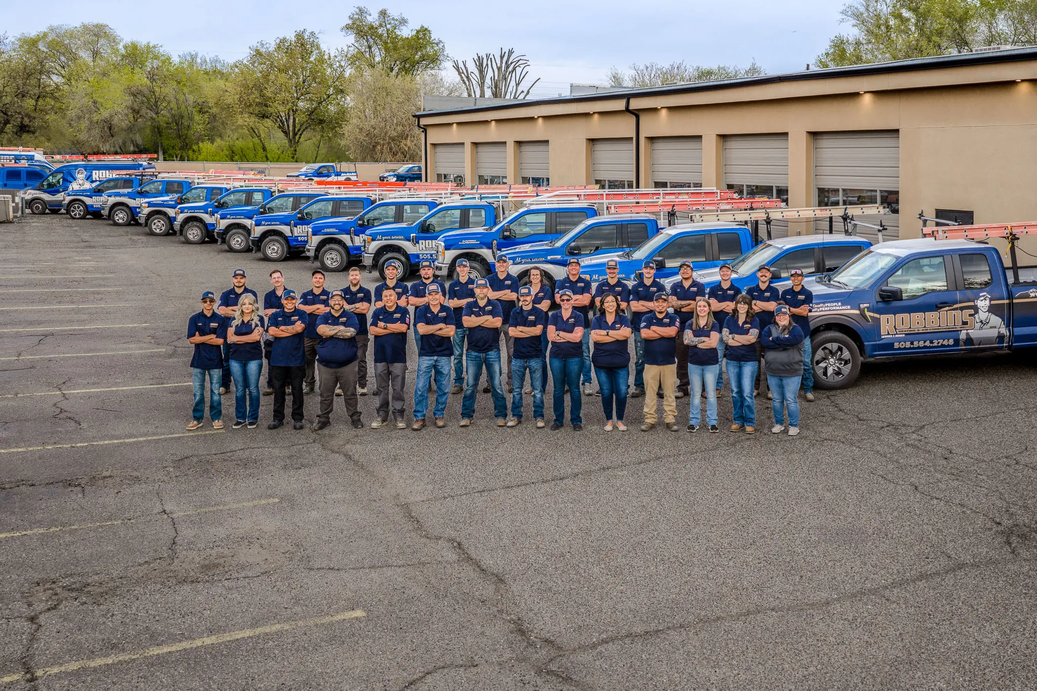 A group of people in matching shirts stands in front of a row of blue company trucks parked outside a garage on a paved lot, with trees and a tan building in the background.